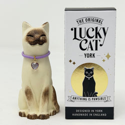 A  Lucky Cat figure hand painted to look like a chocolate point Siamese with a purple collar and heart pendant, shown next to its white and black retail packaging box on a white background.