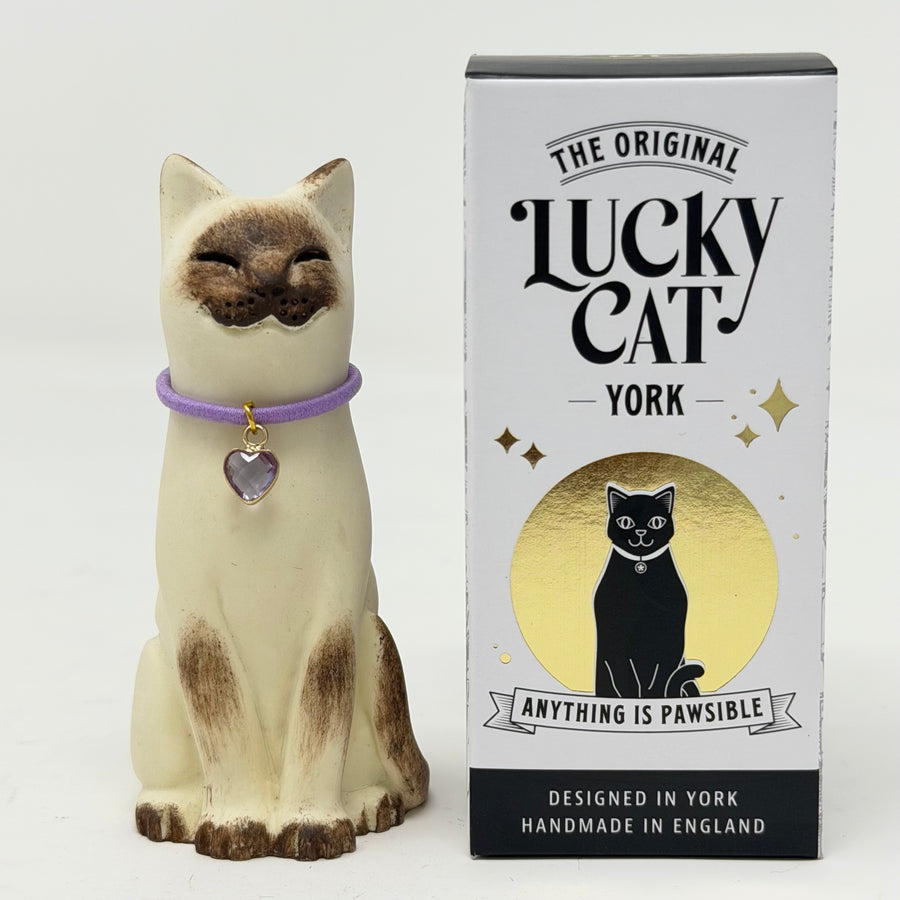 A  Lucky Cat figure hand painted to look like a chocolate point Siamese with a purple collar and heart pendant, shown next to its white and black retail packaging box on a white background.
