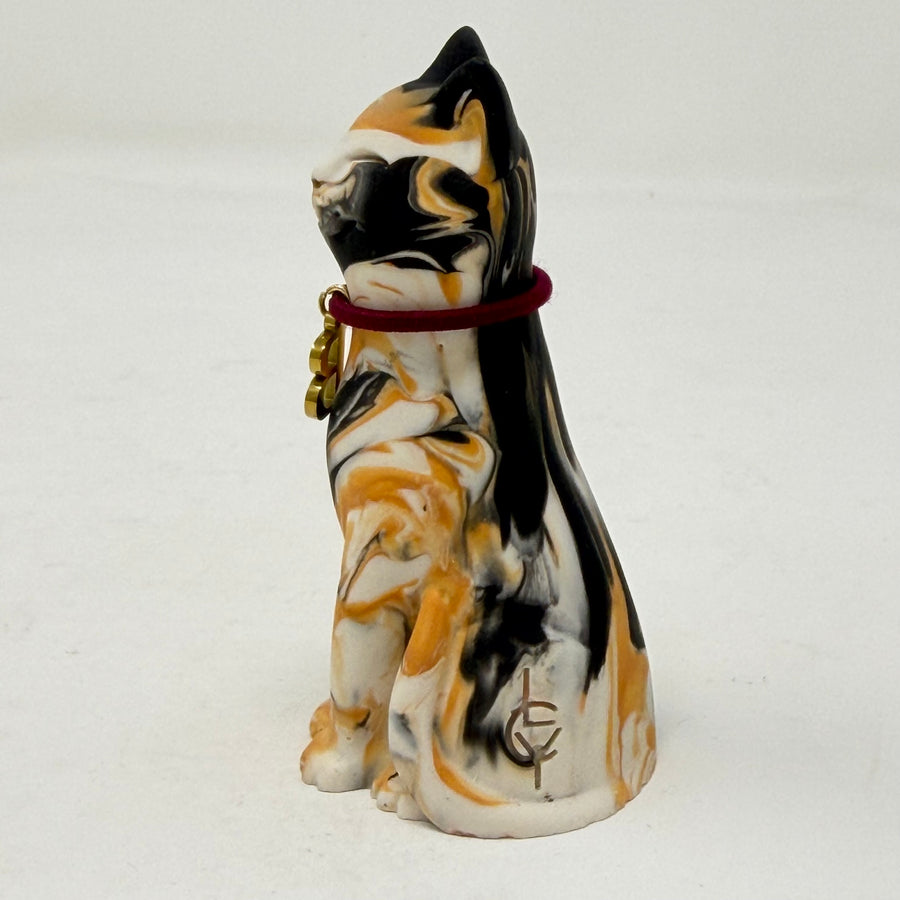 A marbled tortoiseshell Lucky Cat figurine wearing a burgundy collar with a gold letter B charm charm. Side view.