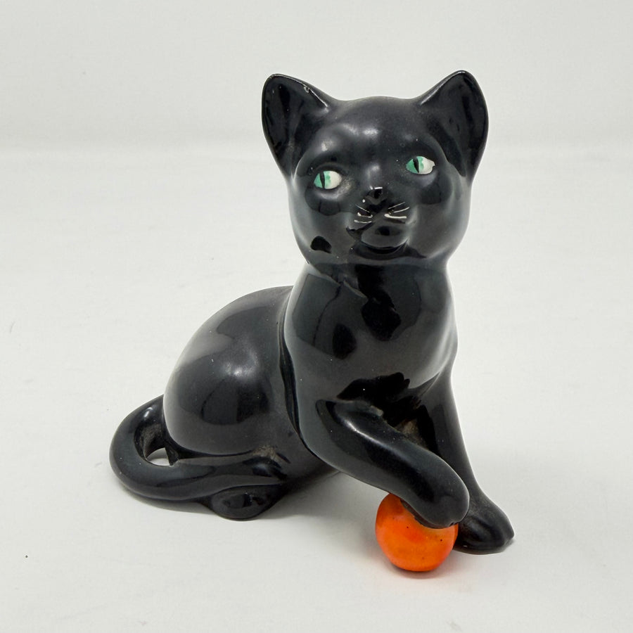 A glossy black ceramic cat figurine sitting and holding a small orange ball, photographed against a plain light background.
