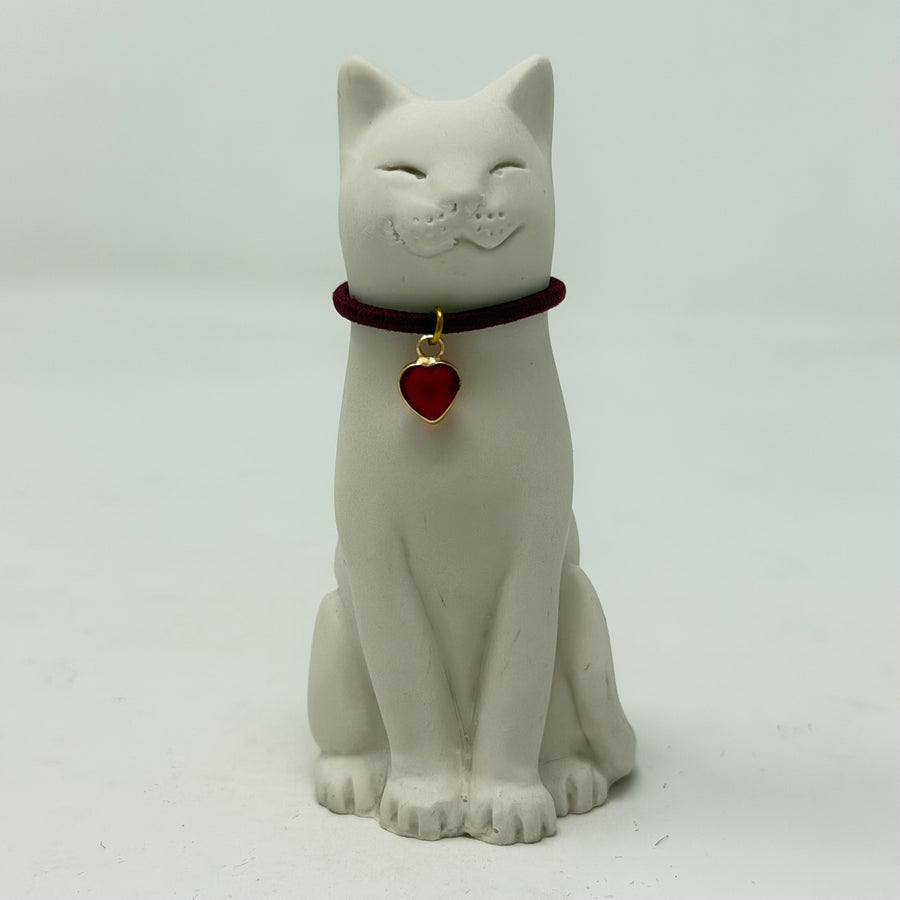 A white resin smiling Lucky Cat figurine wearing a burgundy collar with a red heart charm. Front view.