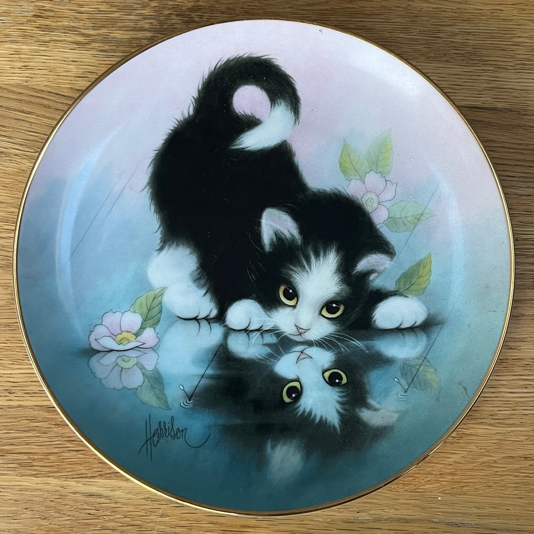 Decorative plate with a painting of a black and white kitten and flowers on a wooden surface.