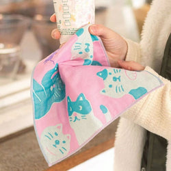 A close-up of a person holding a pink cloth napkin with a playful cat face pattern. The napkin is wiping a paper cup