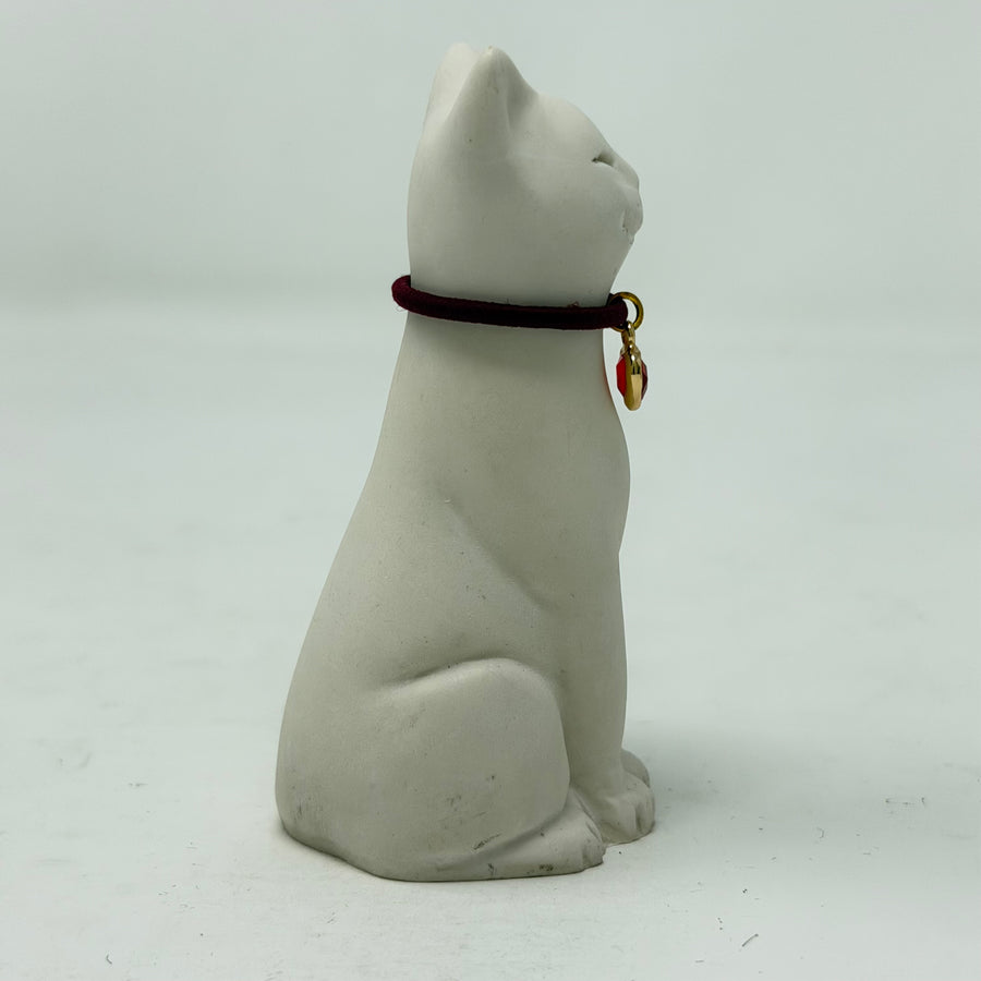 A white resin Lucky Cat figurine wearing a burgundy collar with a red heart charm. Side view.