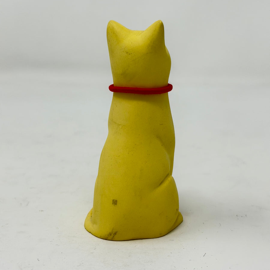 A smiling yellow lucky cat figurine wearing a red collar with a round Union Jack charm, photographed against a plain light grey background - rear view
