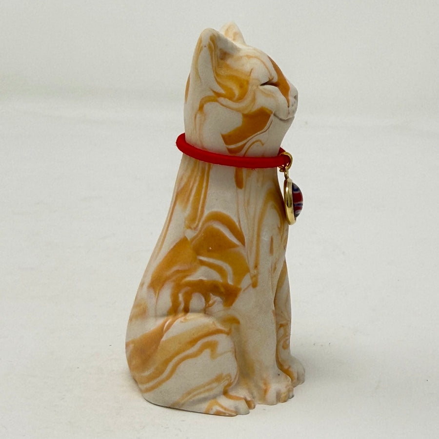 A ginger and white marbled Lucky Cat figurine wearing a red collar with a Union Jack medallion charm. Side view.