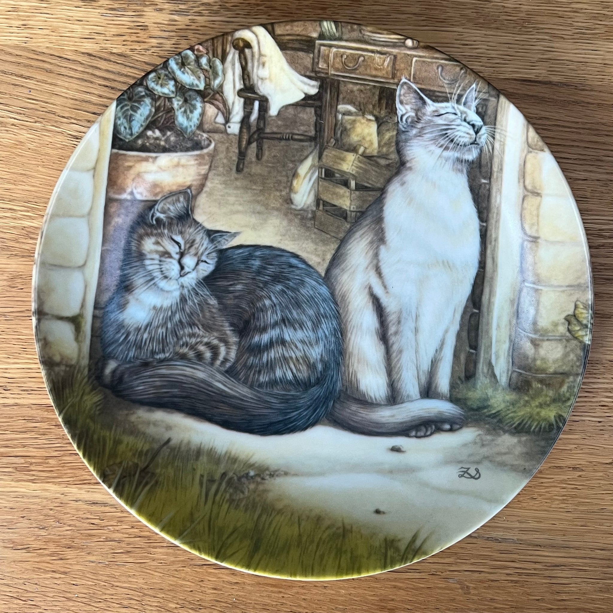 Decorative plate with a painting of two cats in a room setting on a wooden surface.