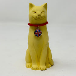 A smiling yellow lucky cat figurine wearing a red collar with a round Union Jack charm, photographed against a plain light gray background.