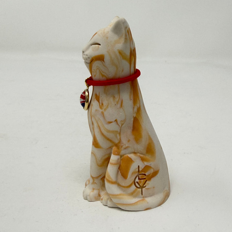 A ginger and white marbled Lucky Cat figurine wearing a red collar with a Union Jack medallion charm. Side view.