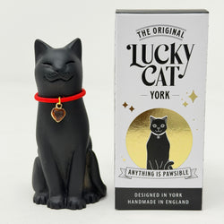 A black cat figurine wearing a bright red collar with a gold heart charm, standing next to its white and gold branded packaging box labeled 'The Original Lucky Cat York'.