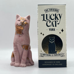 An aubergine coloured Lucky Cat figurine with rose gold foiled accents sits next to its cream-colored packaging box featuring the text 'The Original Lucky Cat York'.