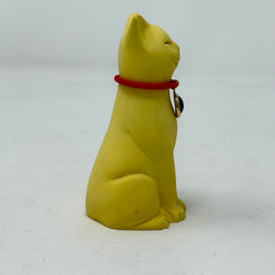A smiling yellow lucky cat figurine wearing a red collar with a round Union Jack charm, photographed against a plain light grey background. Side on view