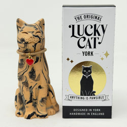 A resin Lucky Cat figurine with a marbled tan and black glaze, wearing a brown rope collar with a red heart charm, shown next to its white and gold branded retail box.