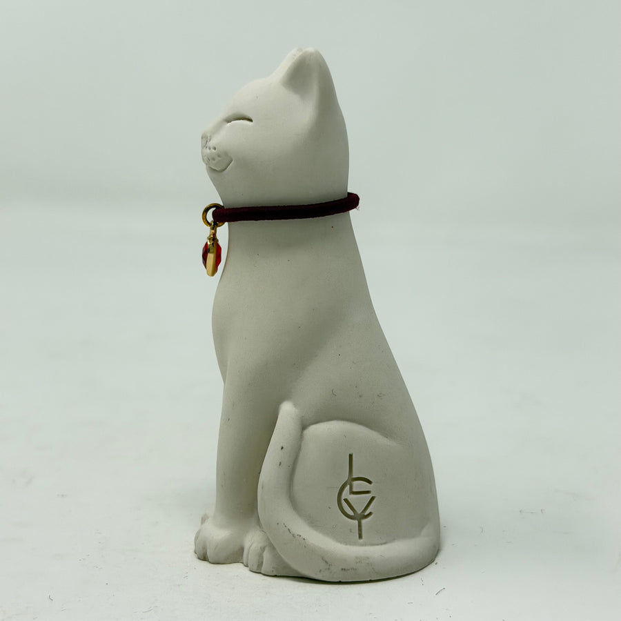 A white resin Lucky Cat figurine wearing a burgundy collar with a red heart charm. Side view.