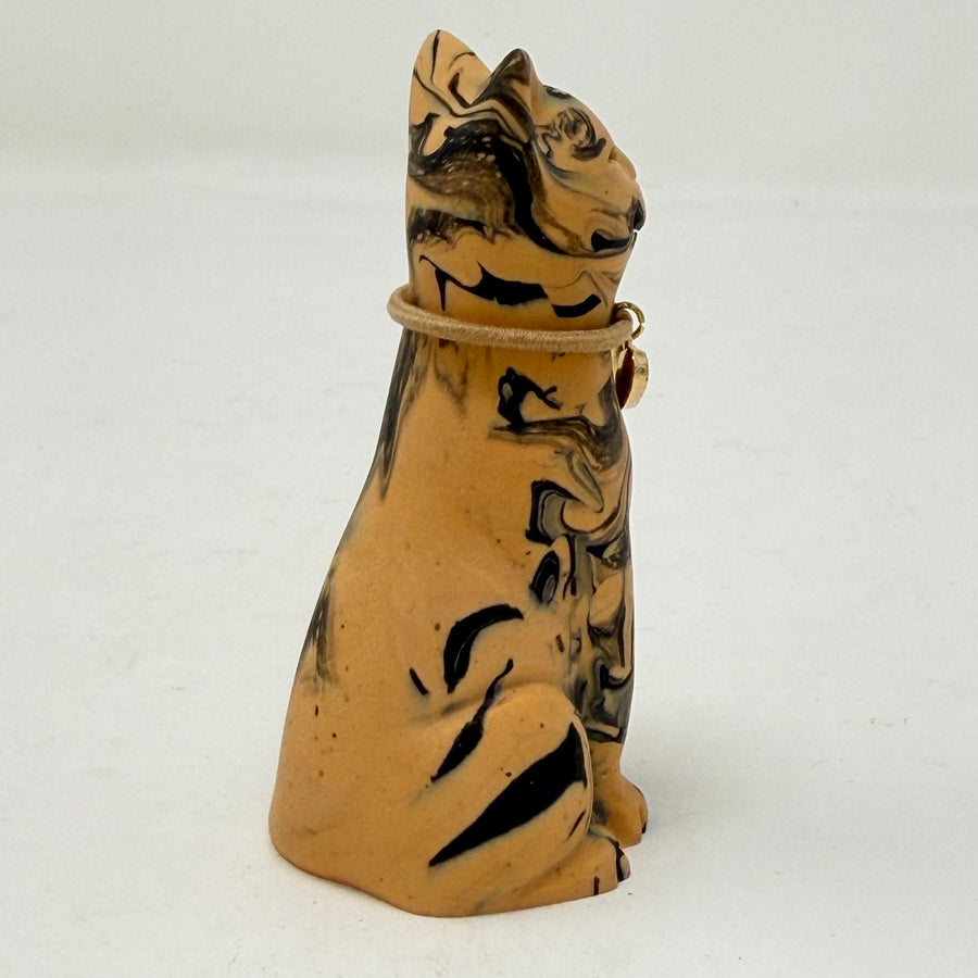 A resin cat figurine with a tan and black marbled pattern, wearing a brown collar with a red heart-shaped charm, set against a plain white background. Side view.