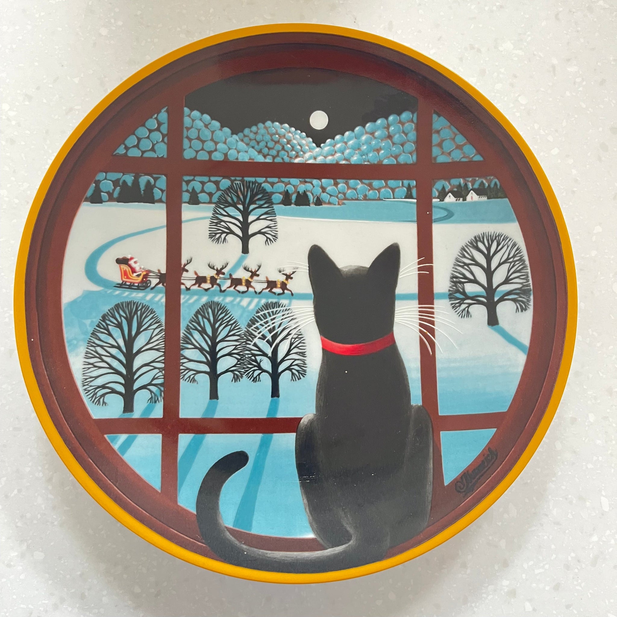 Decorative plate with a cat design looking out a window at trees and snow.