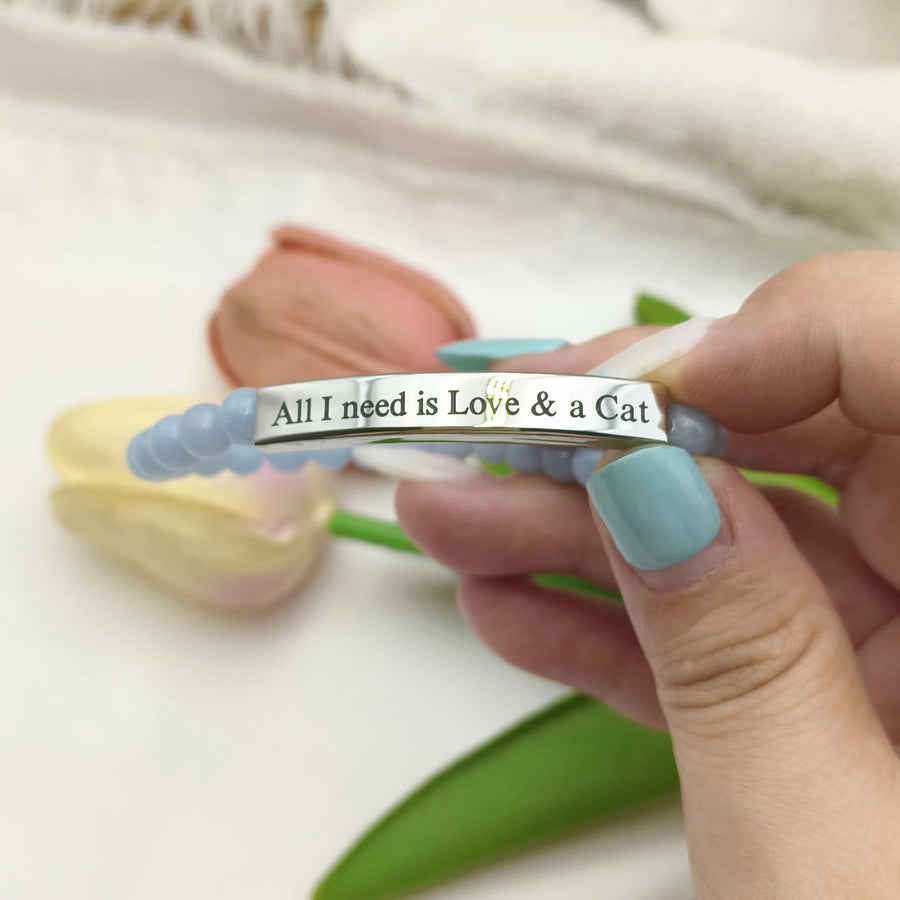 A close-up of a hand with light blue nail polish holding a silver beaded bracelet engraved with 'All I need is Love & a Cat'. 