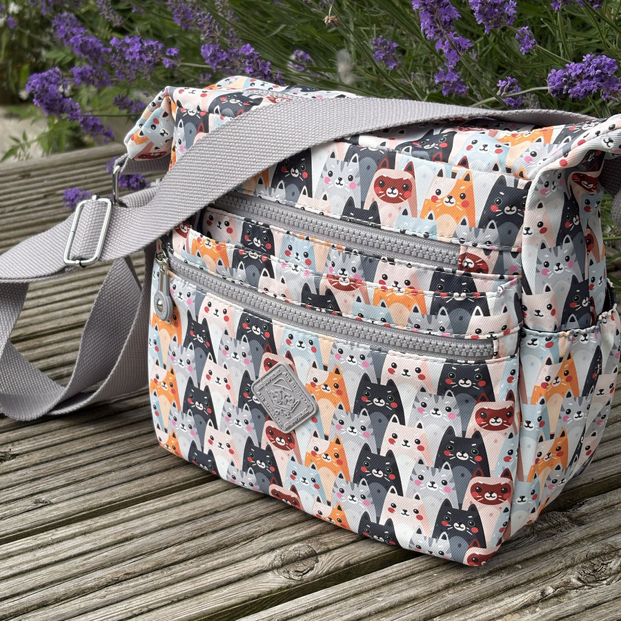 Multicolored cat-patterned bag with a grey strap and two visible zipped pockets on a wooden surface with lavender in the background