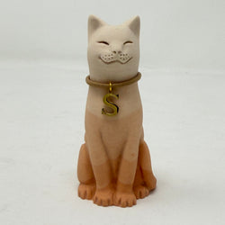 A seated lucky cat figurine in a warm peach-beige tone, wearing a tan collar with a gold initial letter S pendant