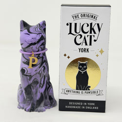 A purple and black marbled cat figurine wearing a purple collar with a gold letter P charm, shown next to its white and black packaging box featuring the text 'The Original Lucky Cat York'.