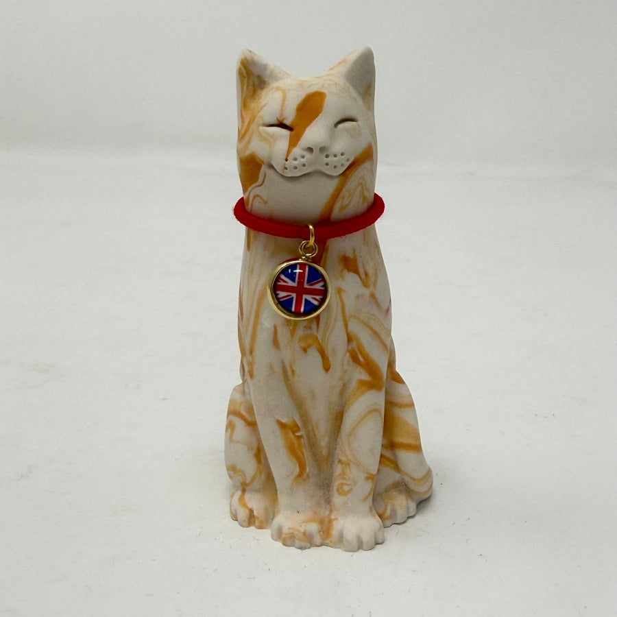 A ginger and white marbled Lucky Cat figurine wearing a red collar with a Union Jack medallion charm. Front view.