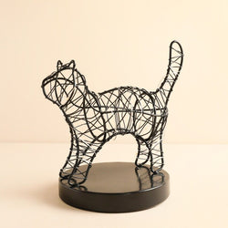 A black wire sculpture of a cat standing on a round black base, photographed against a plain warm beige background.