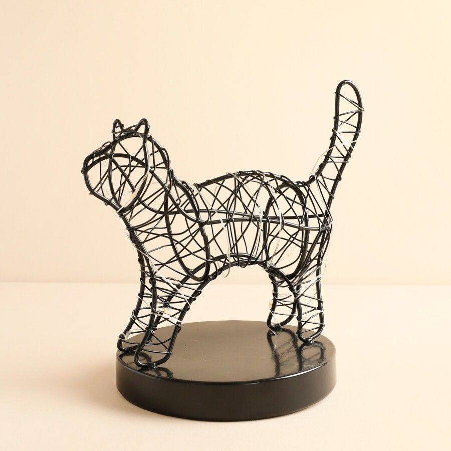 A black wire sculpture of a cat standing on a round black base, photographed against a plain warm beige background.