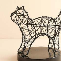 A black wire sculpture of a cat standing on a round black base, photographed against a plain warm beige background.