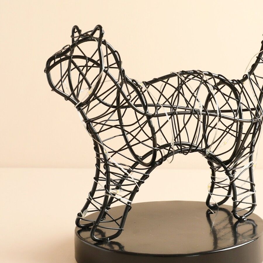 A black wire sculpture of a cat standing on a round black base, photographed against a plain warm beige background.
