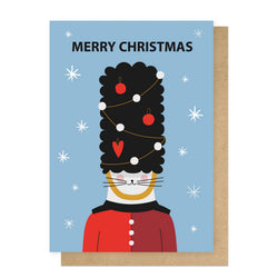 A Christmas greeting card featuring a stylized cat dressed as a toy soldier with a black bearskin hat decorated with ornaments
