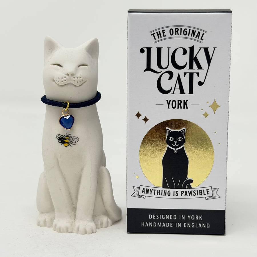 A white resin Lucky Cat figurine wearing a navy blue collar with a blue heart charm and a bee charm, sitting next to its branded box 'The Original Lucky Cat York'
