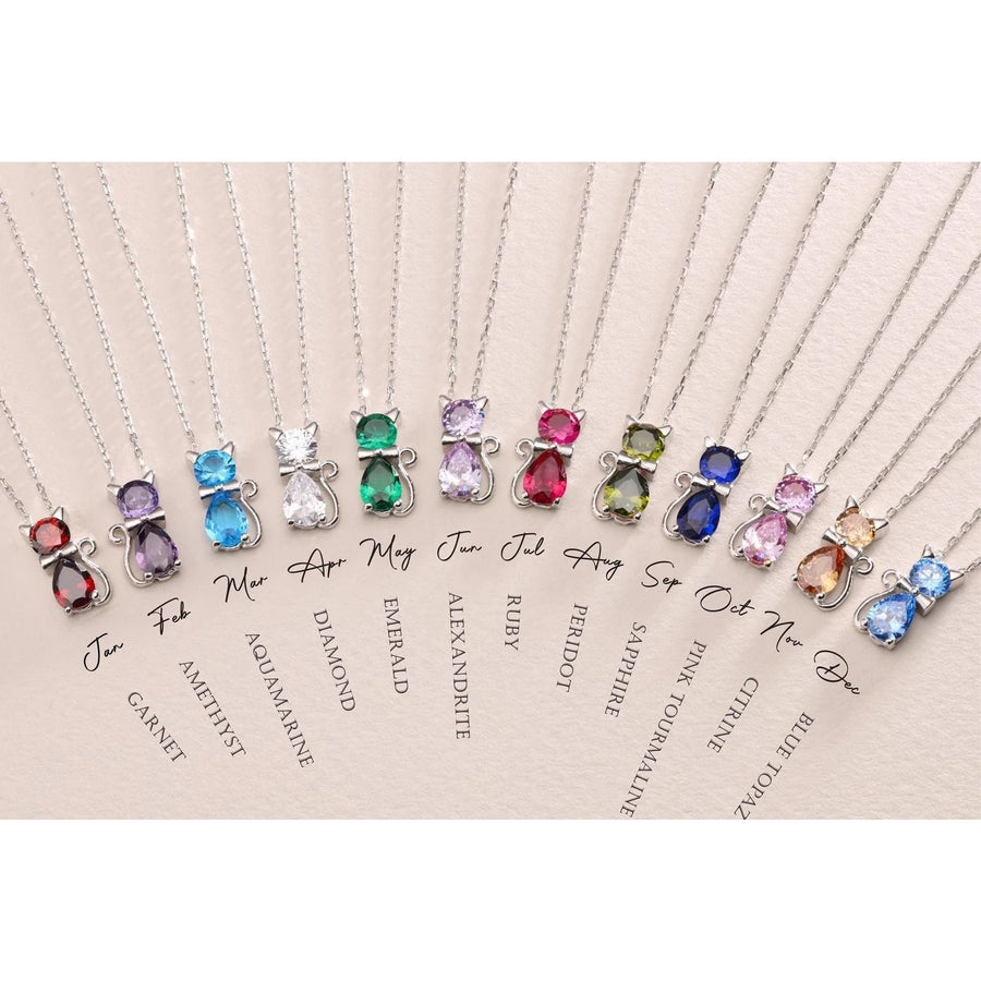 A collection of cat-shaped birthstone pendants on silver chains, each pendant is a different colour representing various birth months