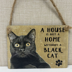 A rectangular hanging sign with a rustic tan background, featuring a black cat illustration and the phrase 'A HOUSE IS NOT A HOME WITHOUT A BLACK CAT