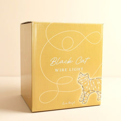 A square cardboard box for the 'Black Cat Wire Light' by Leon Dayal, featuring a mustard yellow cover with white line art of a cat and elegant script text