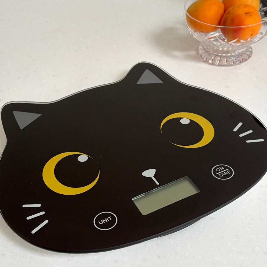 A black digital kitchen scale shaped like a cat face, featuring yellow crescent moon eyes, white whiskers, and a small LCD screen