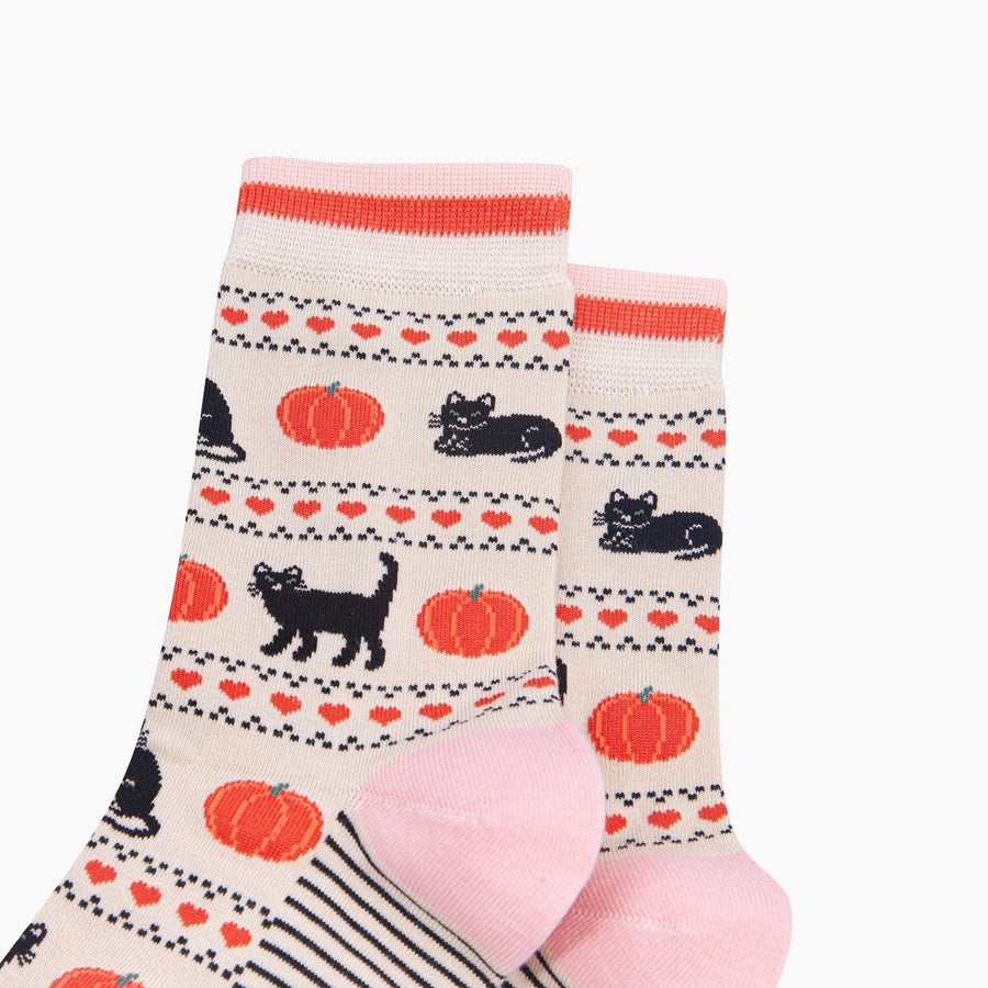 A pair of crew socks with a cozy autumn pattern featuring black cats, orange pumpkins, and red hearts on a cream background