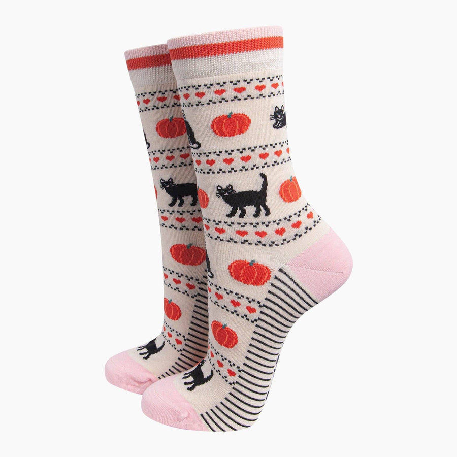 A pair of crew patterned socks, featuring black cats, orange pumpkins, and red hearts on a cream background, with pink toes and heels.