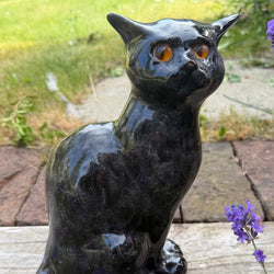 A glossy black cat figurine with orange eyes, sitting on a weathered wooden fence in a garden with purple flowers