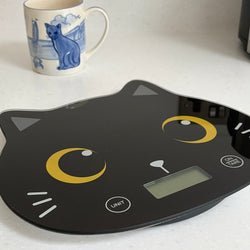 A black cat-shaped digital kitchen scale with yellow crescent eye details, shown next to a blue and white ceramic mug
