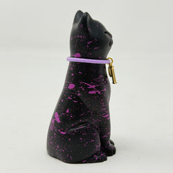 A black lucky cat figurine with vibrant purple paint splatters and a purple collar featuring a small gold charm