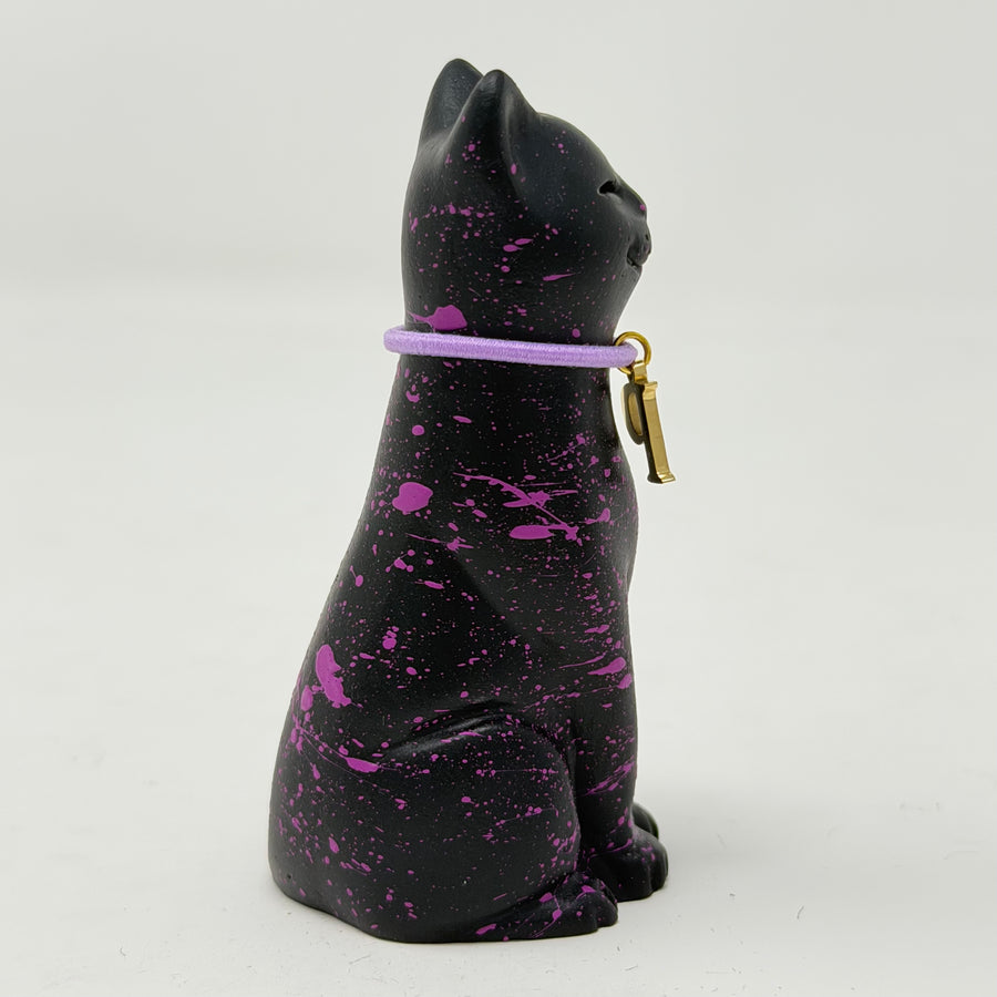 A black lucky cat figurine with vibrant purple paint splatters and a purple collar featuring a small gold charm