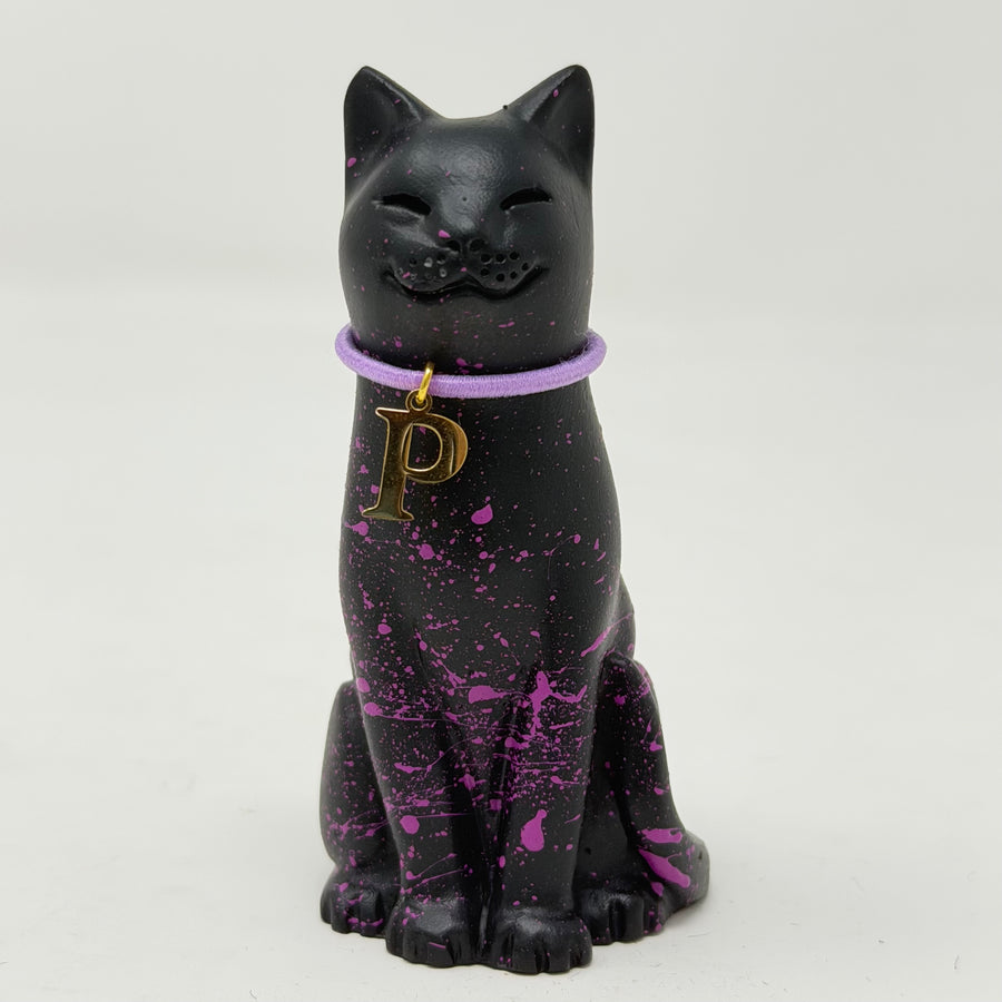 A black lucky cat figurine with purple paint splatters and a purple collar featuring a gold letter 'P' charm, set against a plain light gray background.