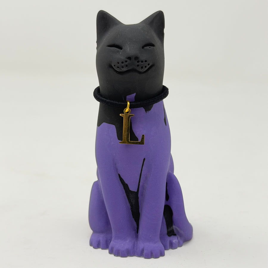 A black and purple lucky cat figurine wearing a black collar with a gold initial 'L' charm