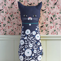A cat-shaped decorative pillow in navy blue fabric with a white floral print on the lower half, featuring a simple embroidered face, set against a pink patterned wallpaper background.