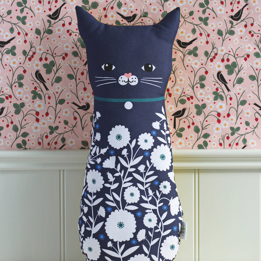 A cat-shaped decorative pillow in navy blue fabric with a white floral print on the lower half, featuring a simple embroidered face, set against a pink patterned wallpaper background.