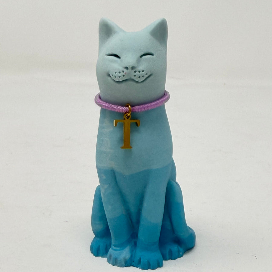 A light blue ombre lucky cat figurine wearing a lavender collar with a gold letter 'T' charm