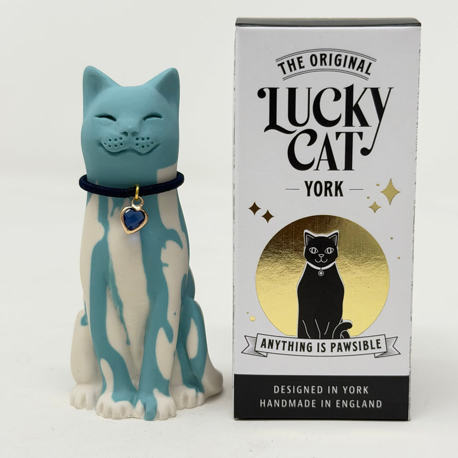 A blue and white lucky cat figurine wearing a black collar with a blue heart charm, displayed next to its white, black, and gold retail box labeled 'The Original Lucky Cat York'.