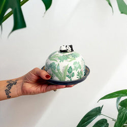 Hand holding a ceramic cheese dome with leaf pattern and cat figurine, surrounded by green leaves