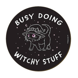A round black coaster with a white line-art illustration of a witch cat wearing a hat, surrounded by the text 'BUSY DOING WITCHY STUFF' 