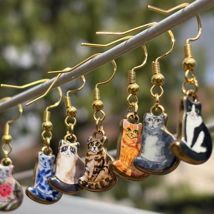 A set of dangling earrings shaped like illustrated cats, hanging from gold hooks. The cat designs include floral, blue-and-white, tabby, orange, gray, and black-and-white patterns